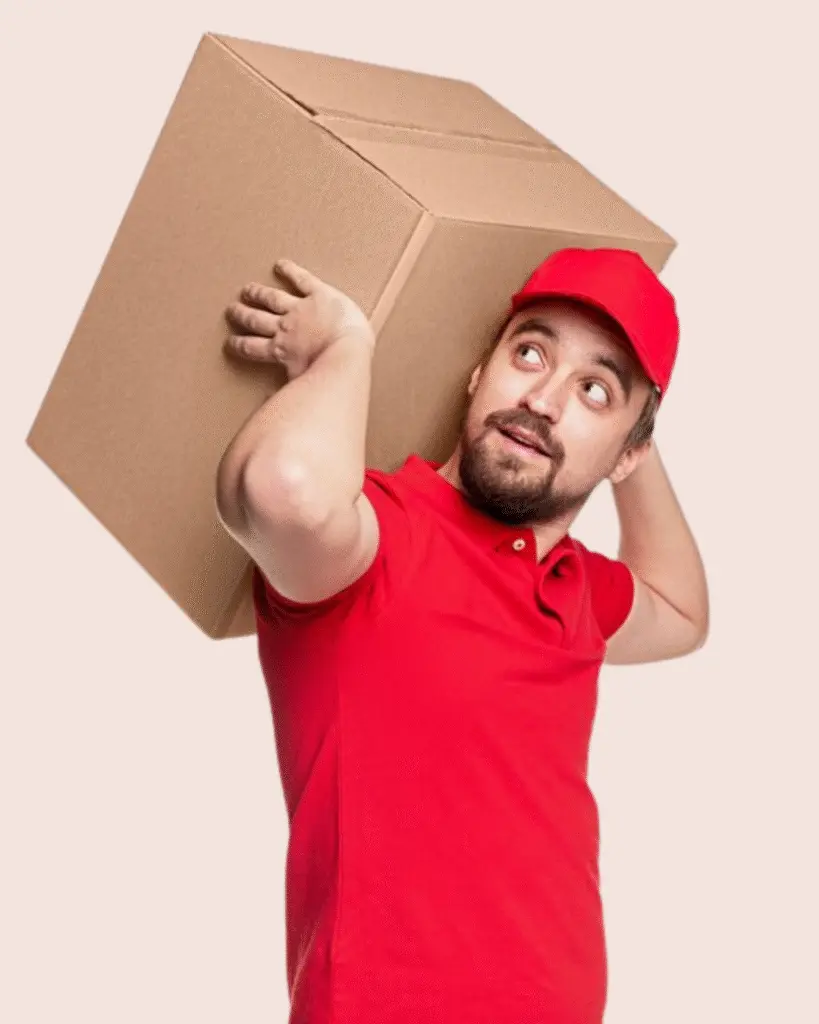 Smiling delivery worker in red uniform holding a large cardboard box on his shoulder with one hand on his hip on a transparent background